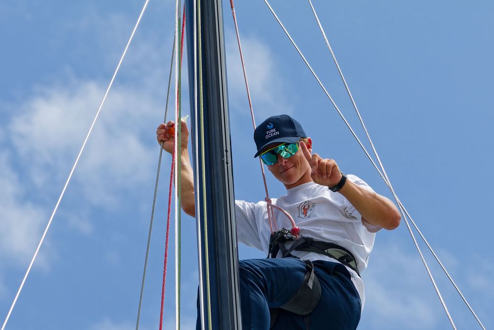A sailor part way up the mast, harnessed on, working the rigging